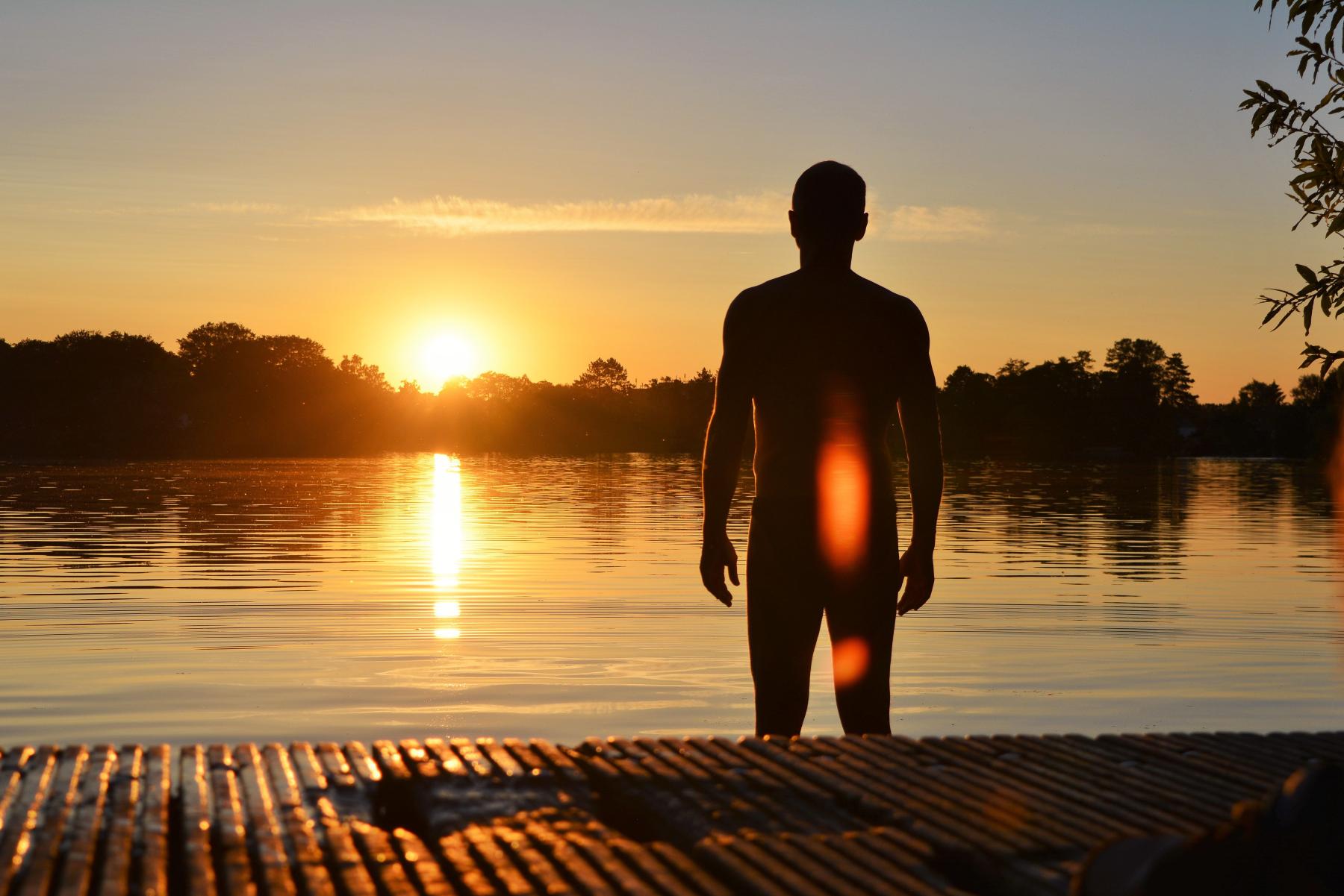 Ein Mann in R&uuml;ckenansicht steht am See und schaut sich den Sonnenuntergang an